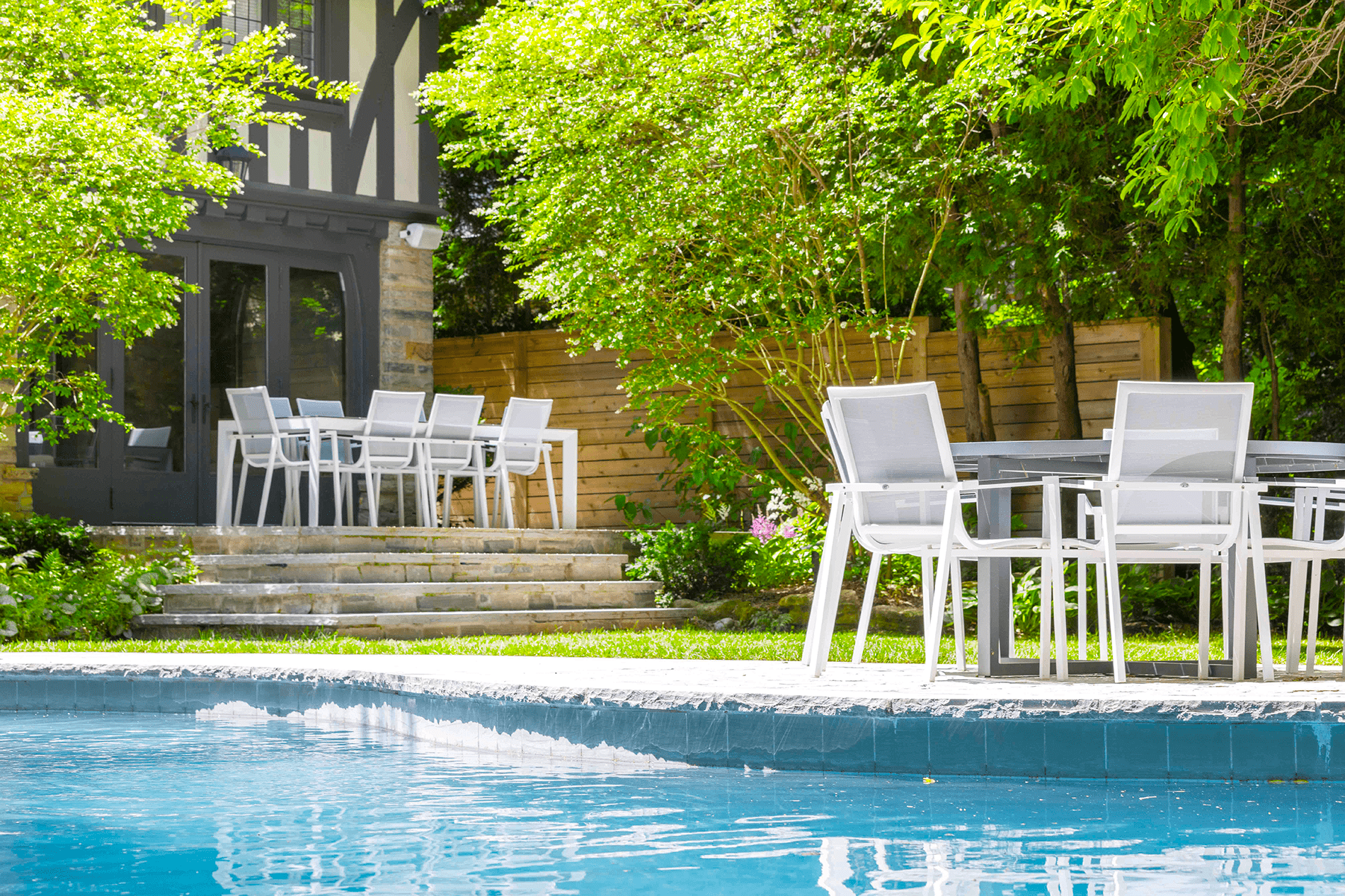 A serene backyard setting with a swimming pool in the foreground and a dining table with chairs near a modern house amidst lush greenery.