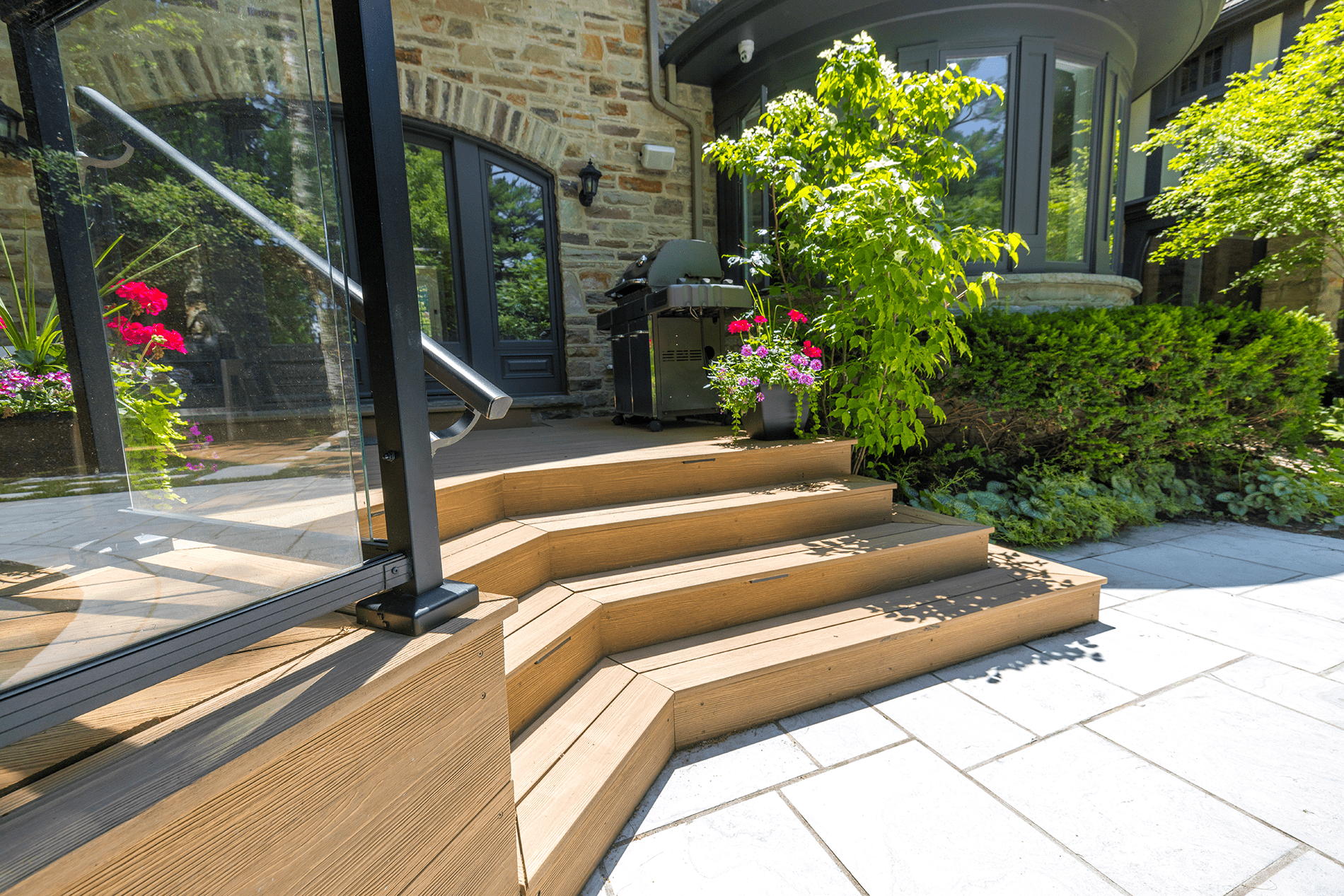 This image displays a modern patio with wooden steps, glass railing, a barbecue grill, vibrant flowers, and part of a stone house with dark windows.