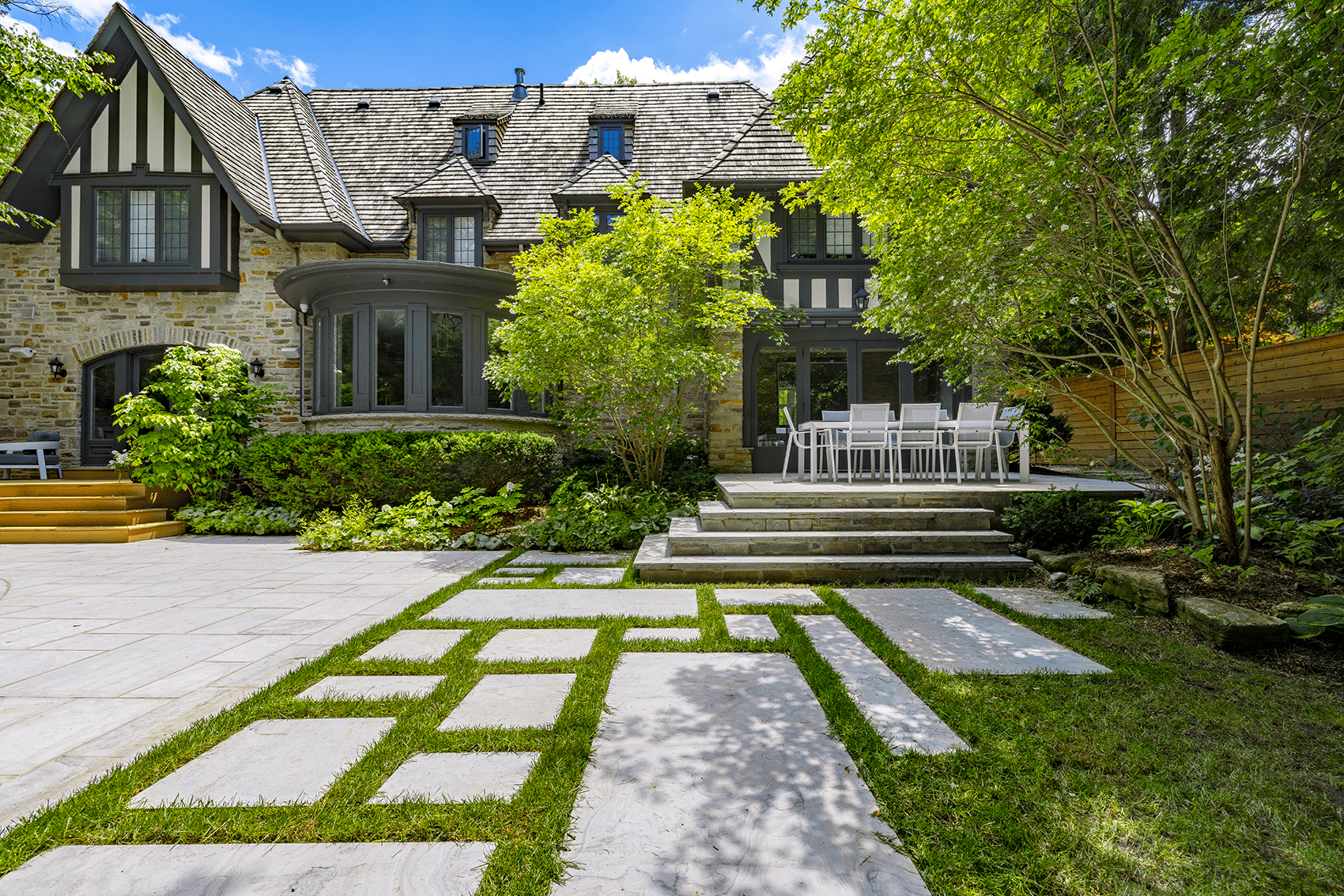 An elegant stone house with timber accents features a manicured lawn, geometric walkway, outdoor dining setup, lush greenery, and a clear blue sky.