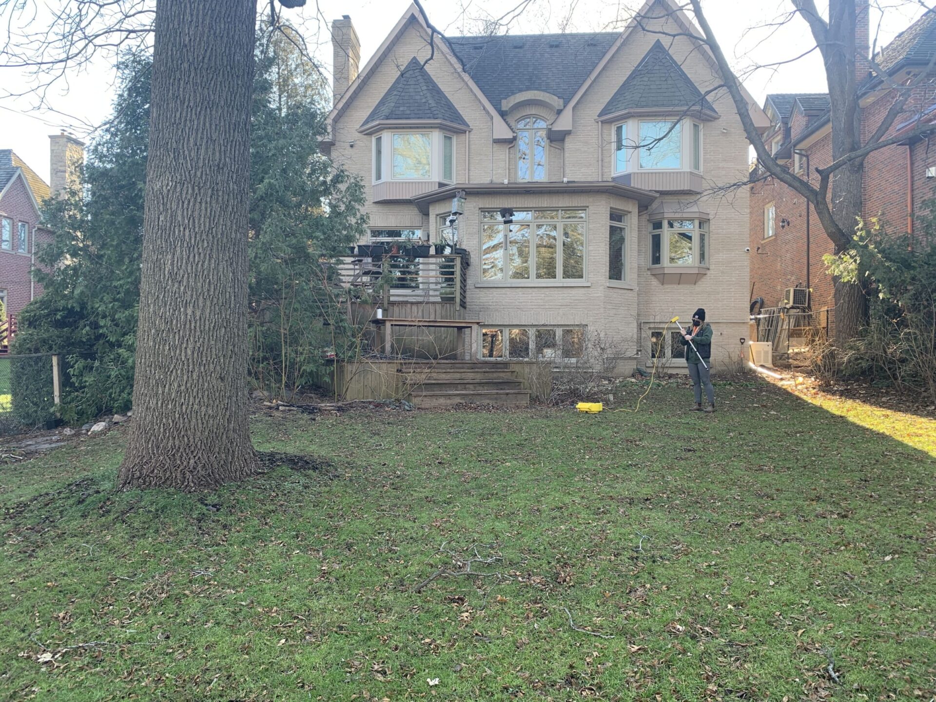 A person rakes leaves in a backyard with sparse trees in front of a large two-story brick house under a clear sky.