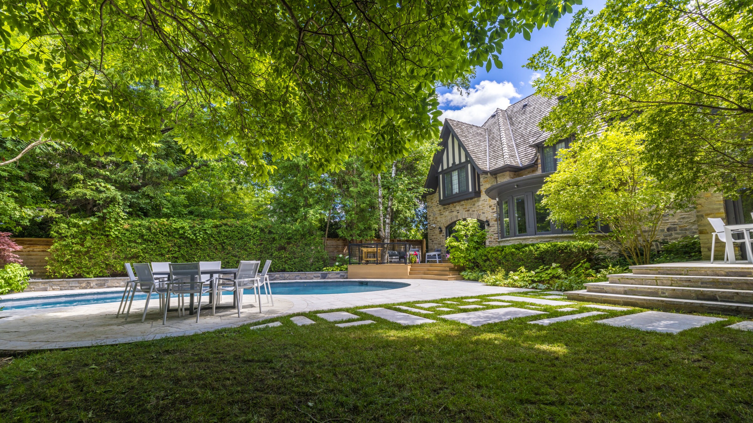 Lush garden with green foliage framing a stone house, swimming pool, patio with dining table and chairs, under a clear blue sky.