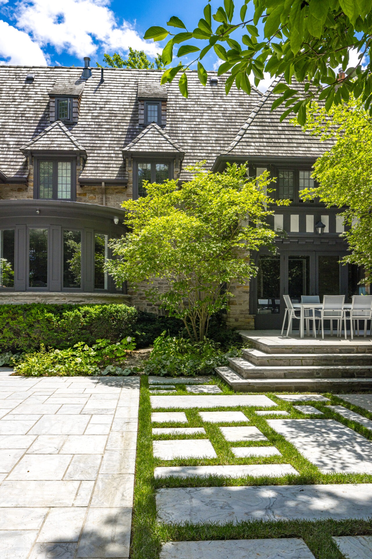 A large stone house with a patterned shingle roof and dormer windows. A manicured lawn, stepping stones, and patio furniture complete the elegant outdoor space.