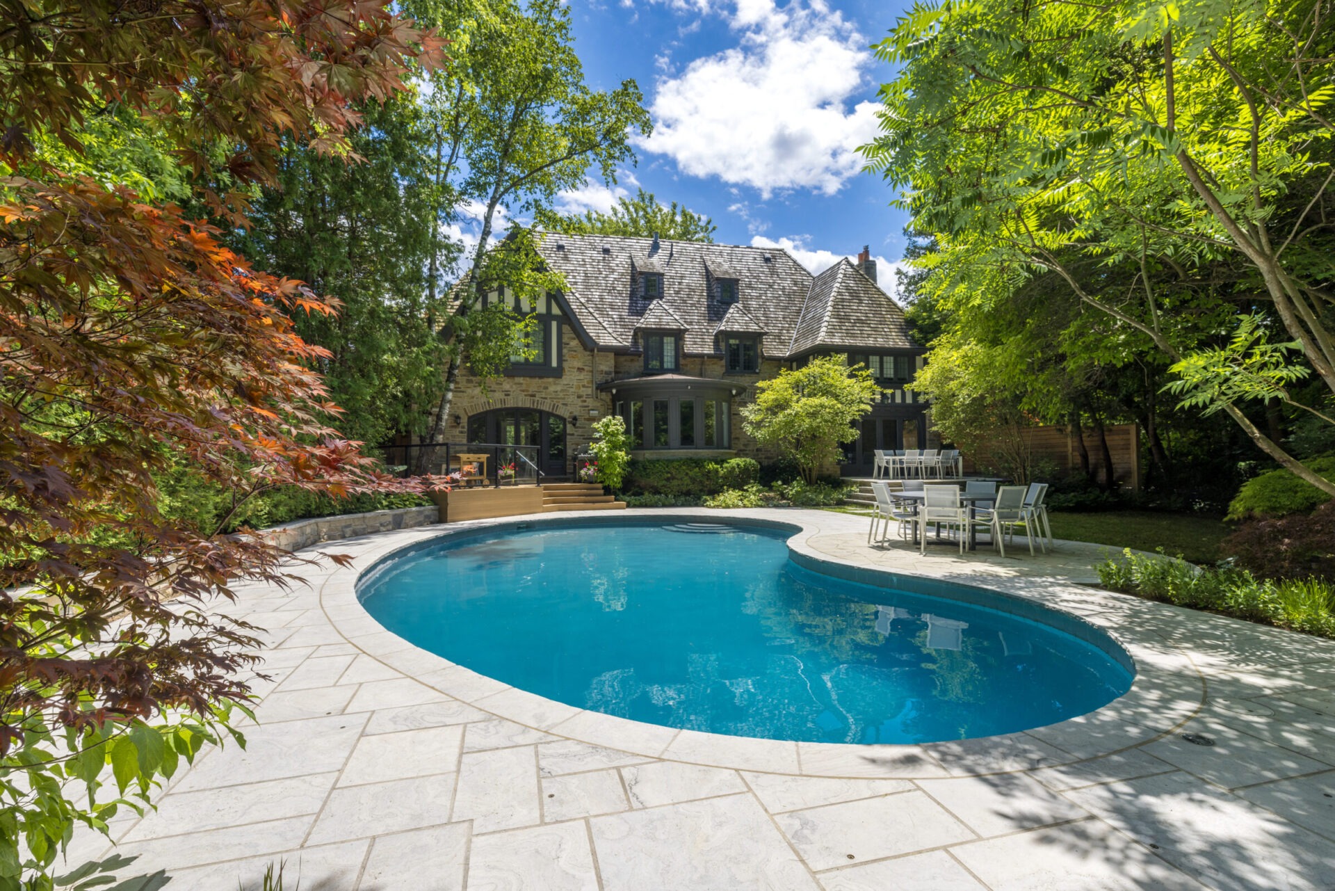 A serene backyard featuring an oval-shaped swimming pool, elegant stone house with a gabled roof, patio furniture, surrounded by lush greenery and trees.