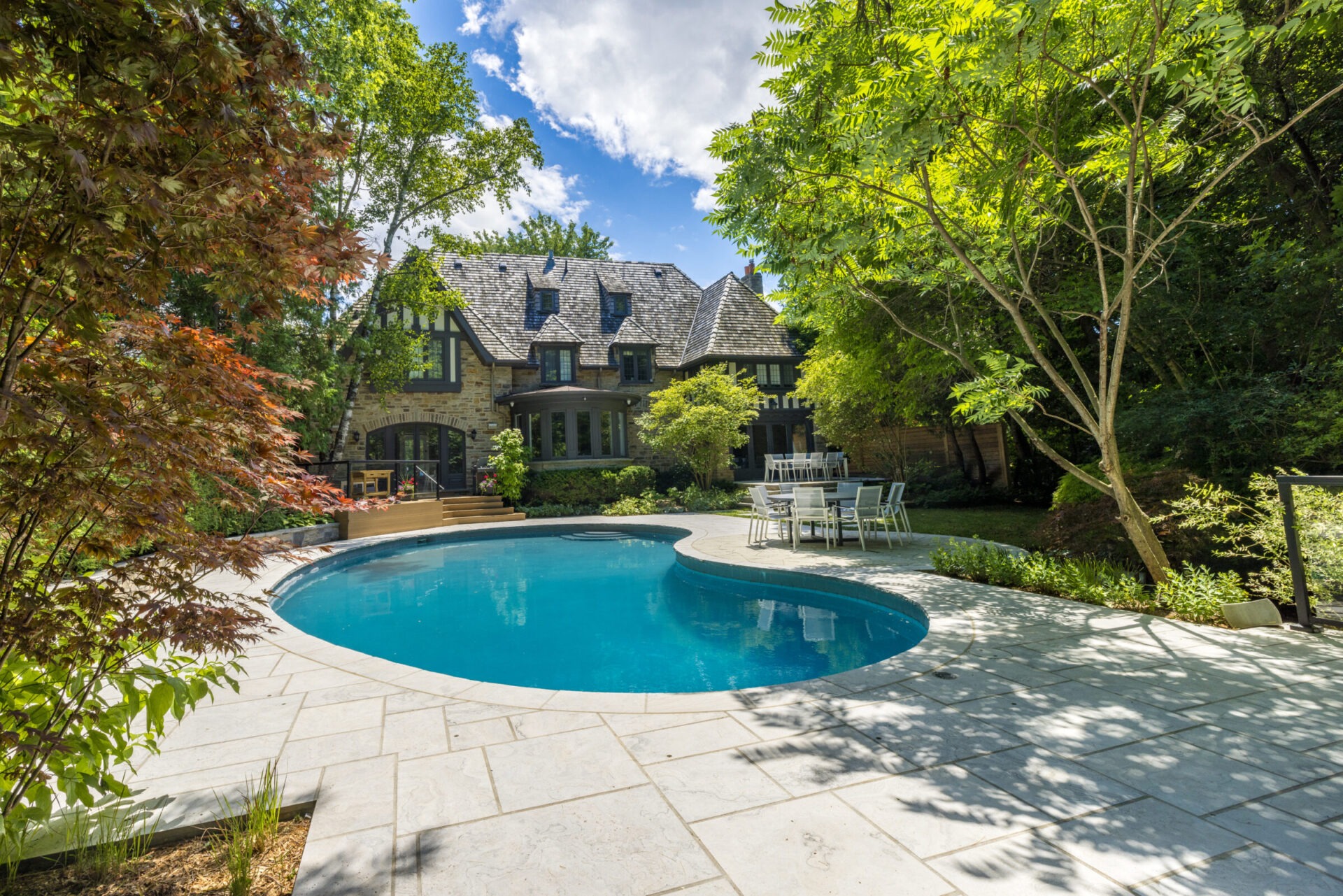 A luxurious stone house with a slate roof, surrounded by verdant trees, overlooks an inviting kidney-shaped swimming pool with a patio and outdoor furniture.