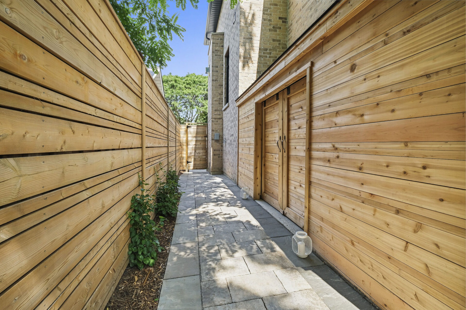 The image depicts a narrow walkway flanked by wooden fences and walls. There are two wooden doors on one side, plants along the path, and a clear sky.