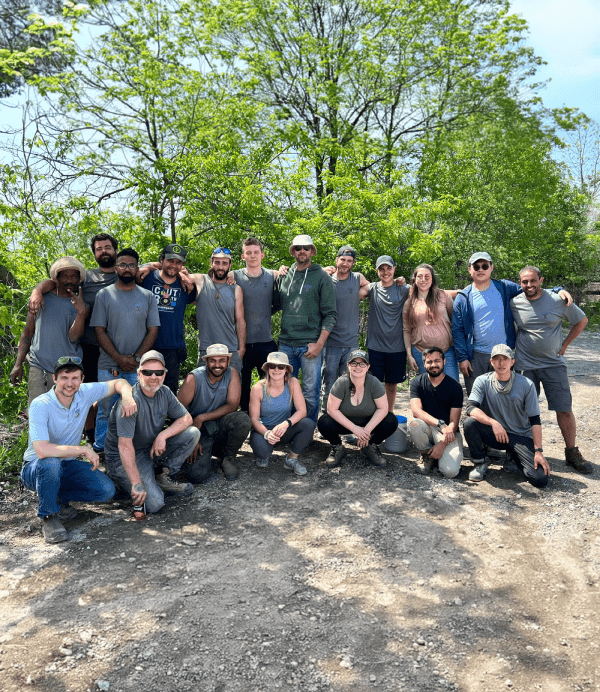 A group of people are posing for a photo outdoors, with trees in the background. They wear casual clothes, and some have hats on.