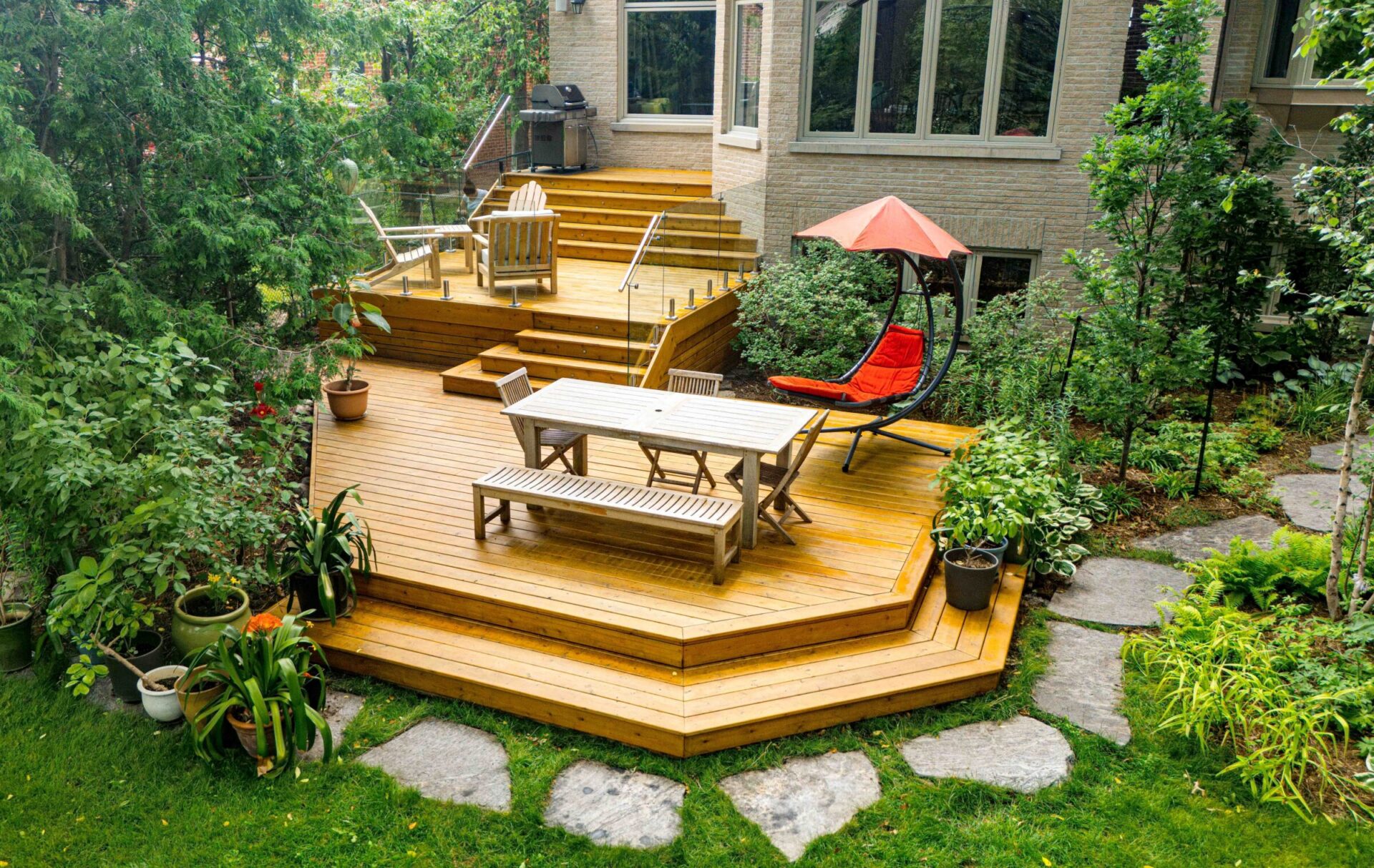 Multi-level cedar deck with glass railings, outdoor dining set, red hanging lounge chair, potted plants, and a built-in BBQ area, surrounded by lush greenery and natural stone garden paths.