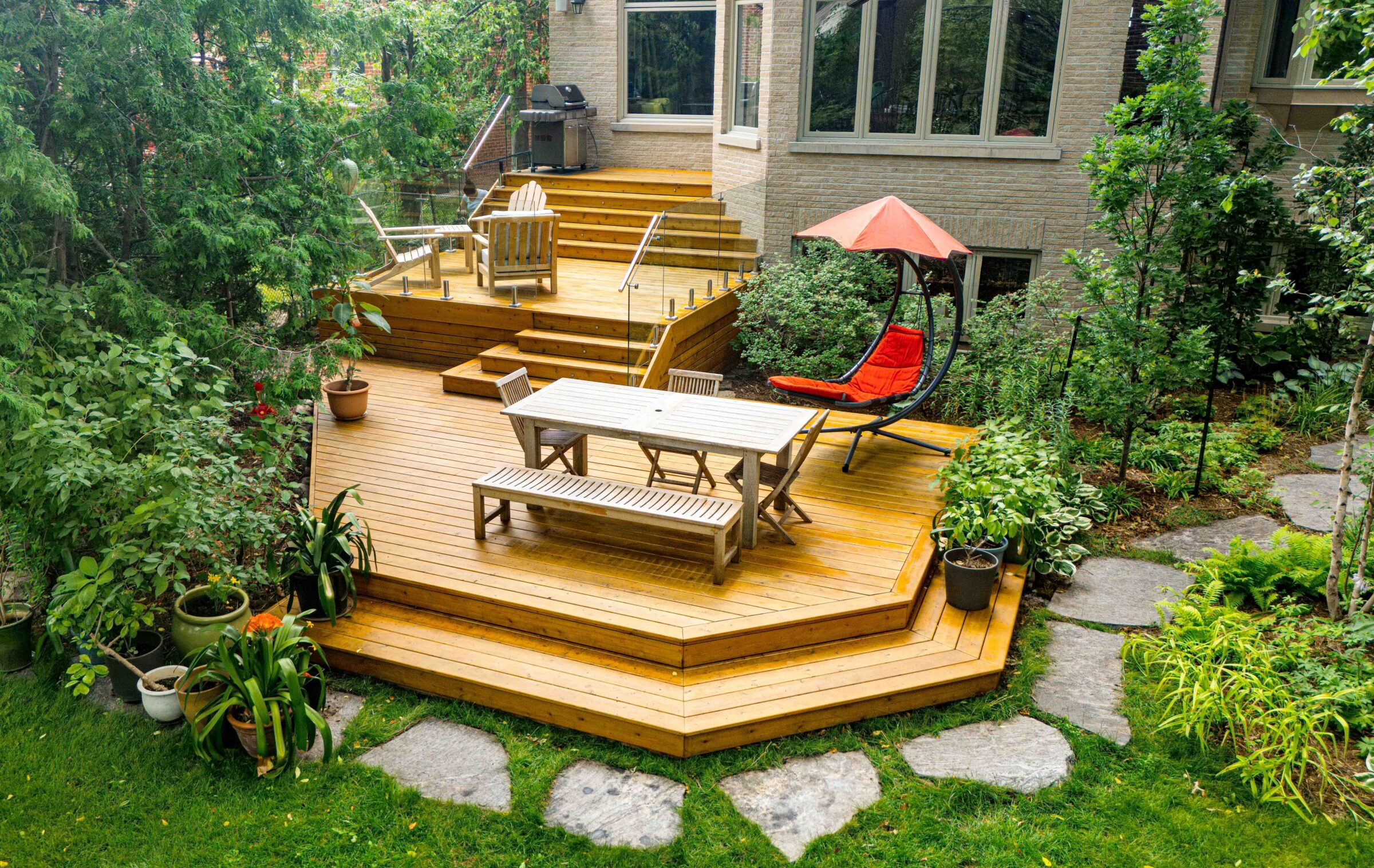 Multi-level cedar deck with glass railings, outdoor dining set, red hanging lounge chair, potted plants, and a built-in BBQ area, surrounded by lush greenery and natural stone garden paths.