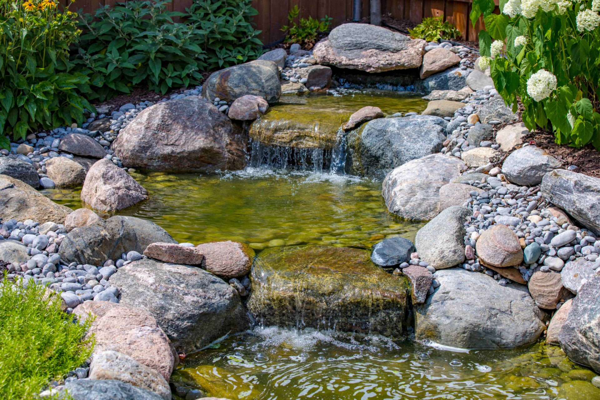 Natural-style backyard water feature with cascading waterfalls over large decorative stones, surrounded by flowering plants, green foliage, and a wooden privacy fence.