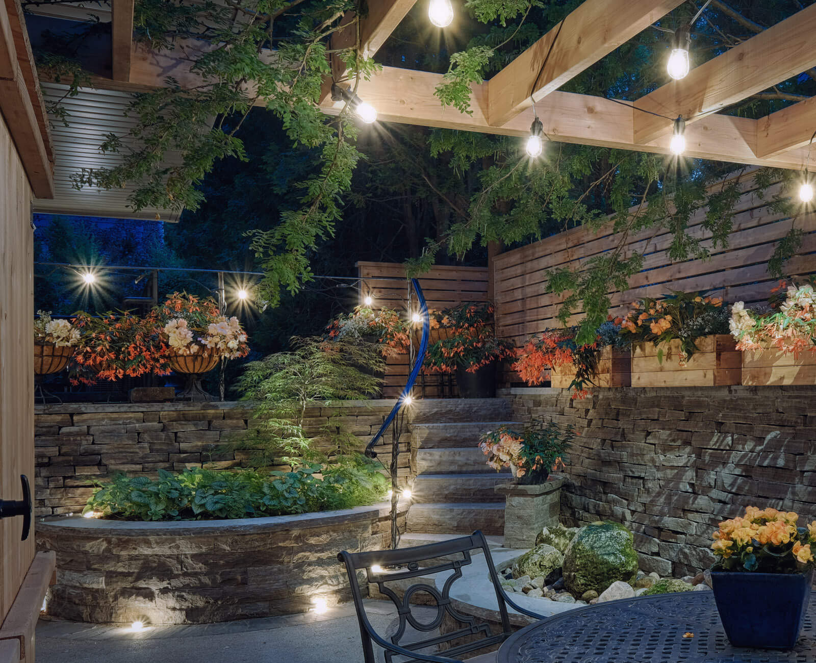 Cozy backyard patio with stone retaining walls, string lights, a pergola, and a round wrought-iron table, surrounded by lush planters and garden steps.