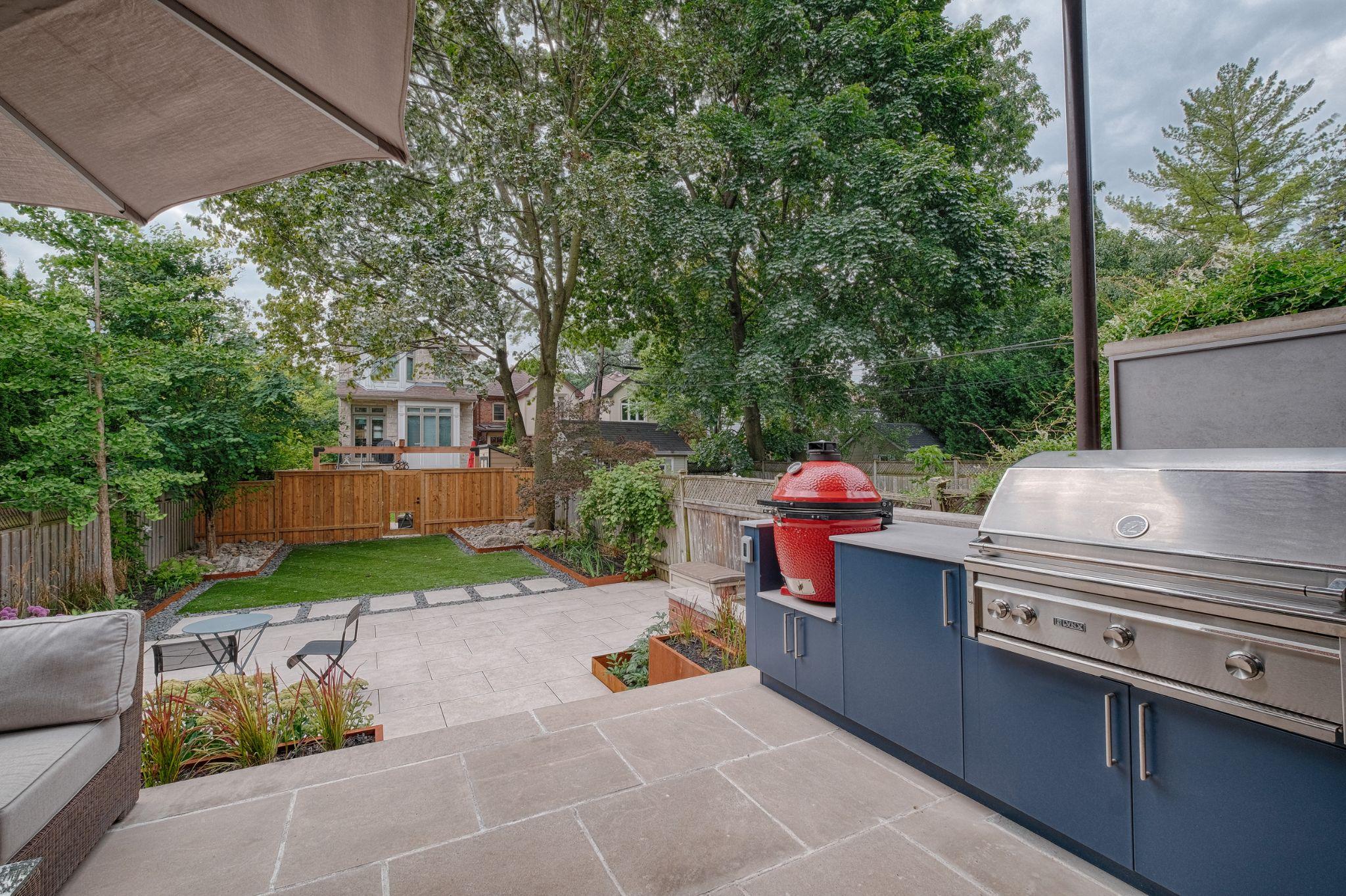 Urban backyard featuring stone patio and outdoor kitchen that includes kamado grill, built-in gas grill, and modern cabinetry. Manicured lawn is located beyond the patio.
