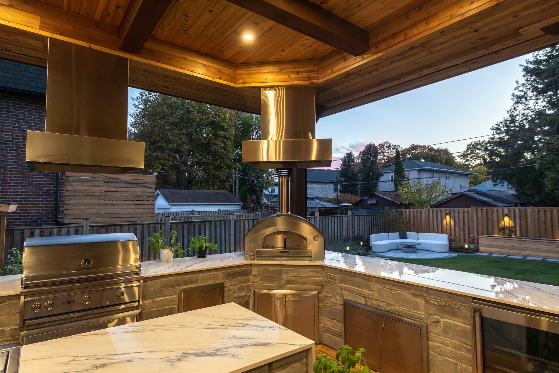 Covered patio outdoor kitchen design with stone base, marble counter, and stainless steel appliances in backyard.