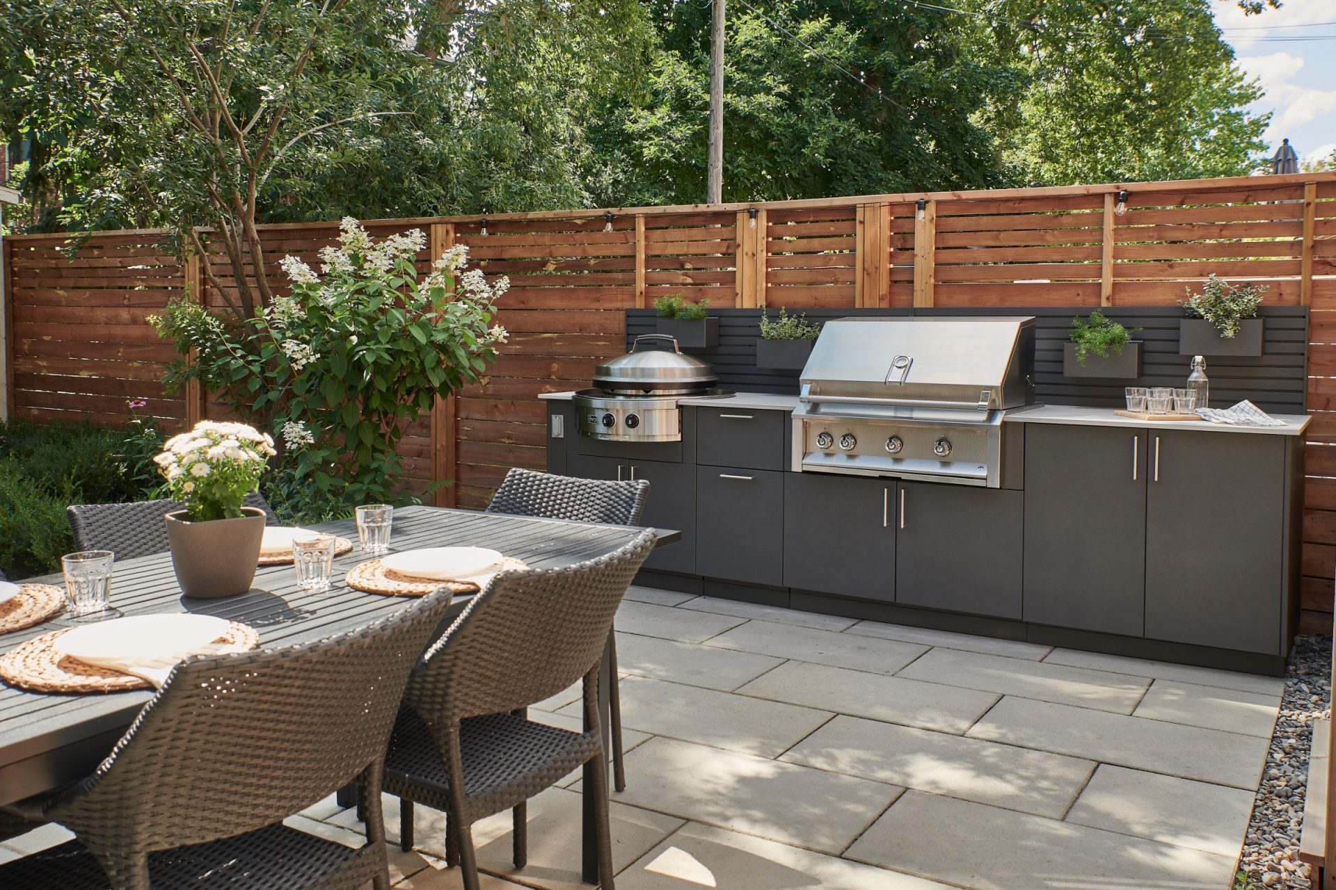 An outdoor kitchen area with stainless steel grill and barbecue sits along a wooden fence in a modern-looking backyard.
